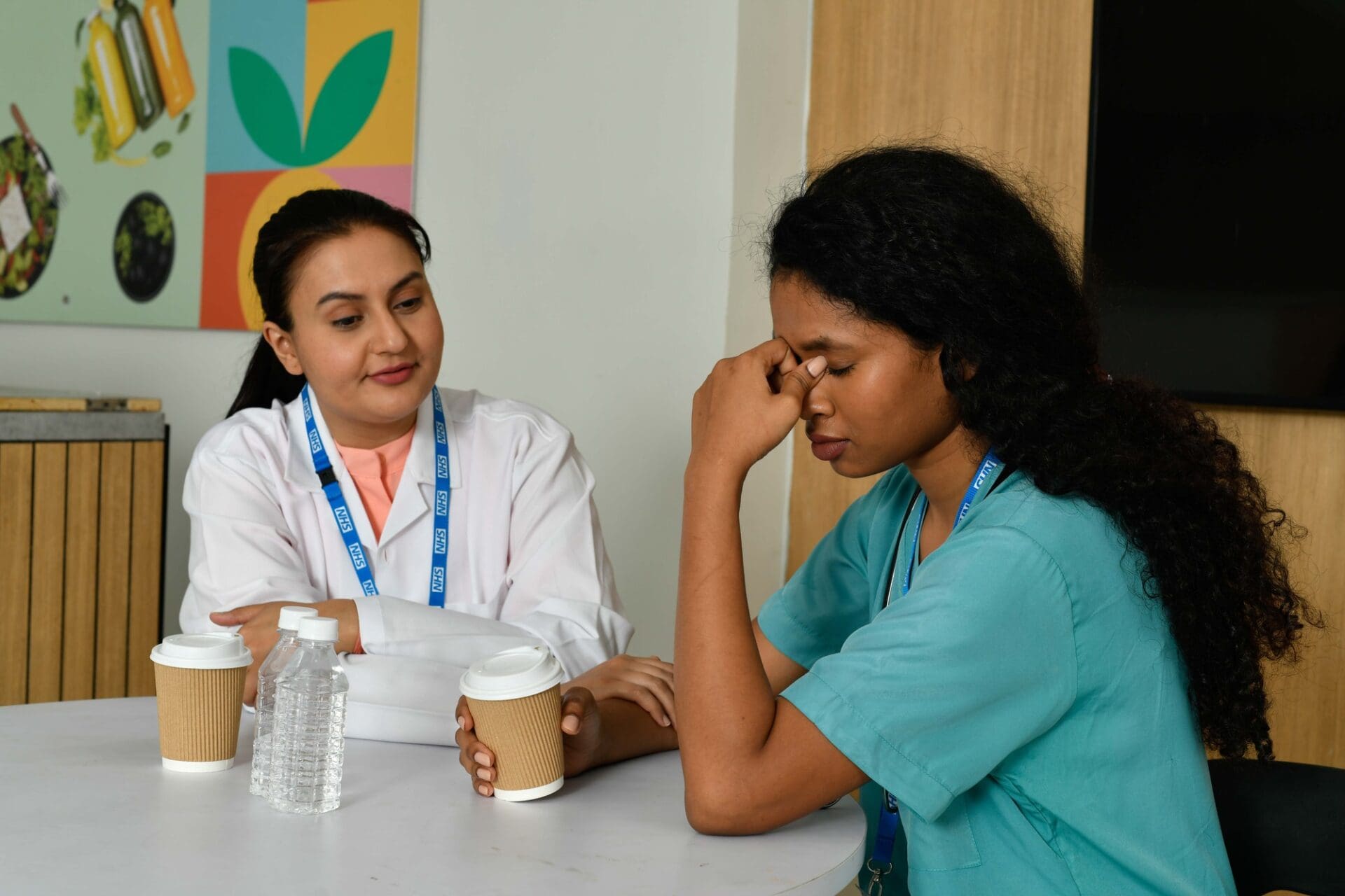 Two doctors sitting at a a table, one with their hand to their face and the other offering support